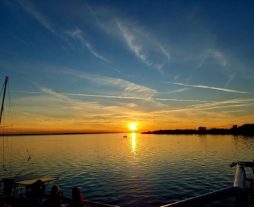 Spokojny zach&oacute;d słońca nad wodą, z delikatnym zarysem łodzi i portowego klimatu po bokach kadru.